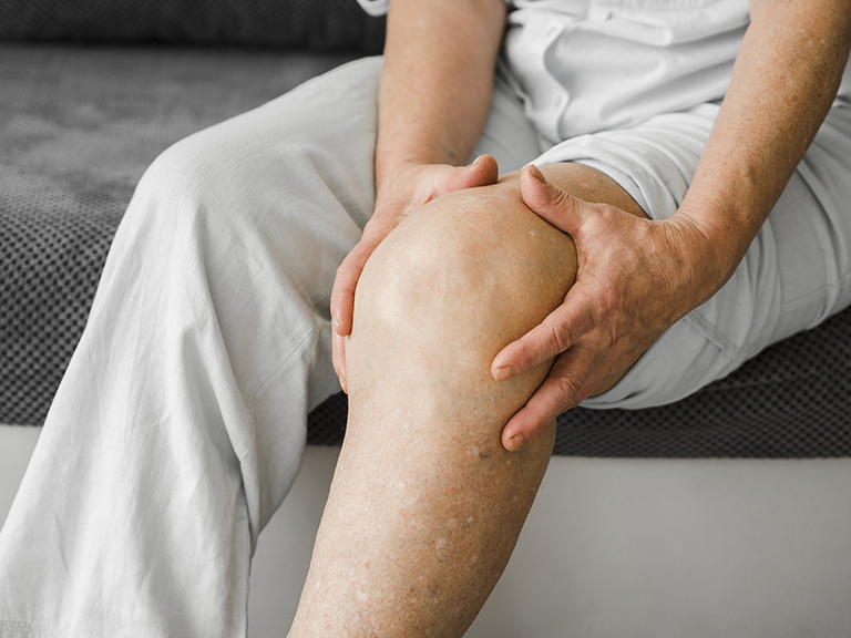 Homem de meia idade com roupa branca, sentado em um sofá preto, aparentando dor no joelho.
