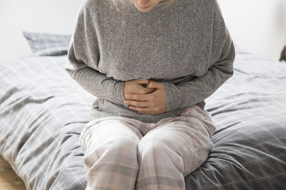 Mulher de meia idade usando blusa cinza e calça quadriculada, sentada na cama segurando suas mãos contra sua barriga em referência a dor abdominal